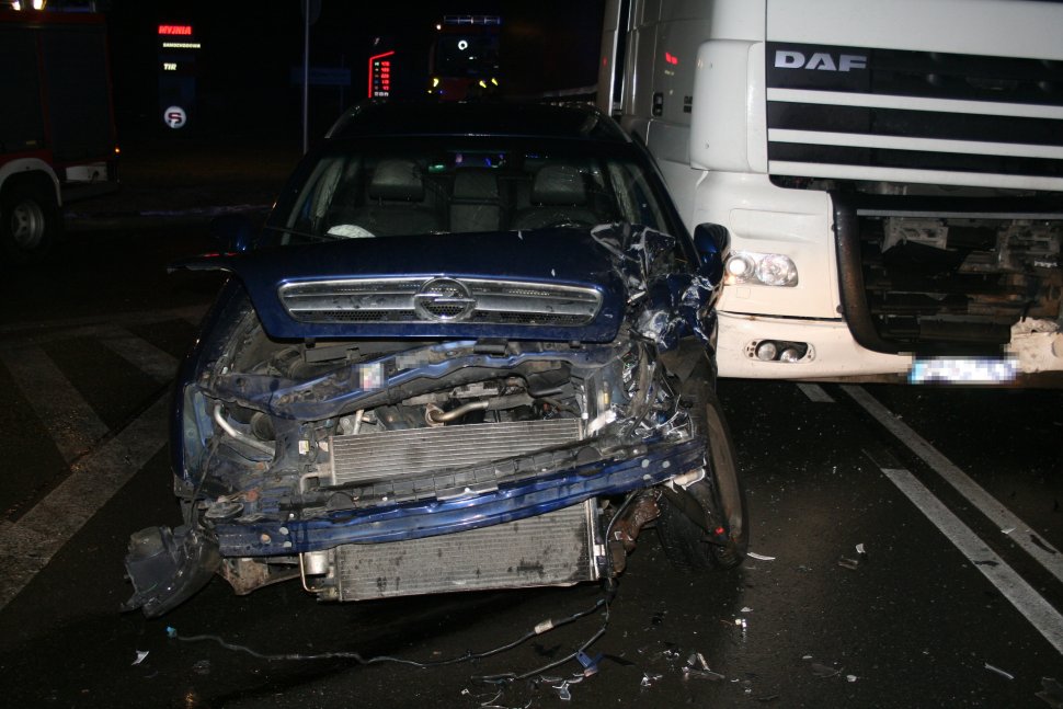z lewej strony fotografii widać przód rozbitego samochodu osobowego, obok którego stoi samochód ciężarowy