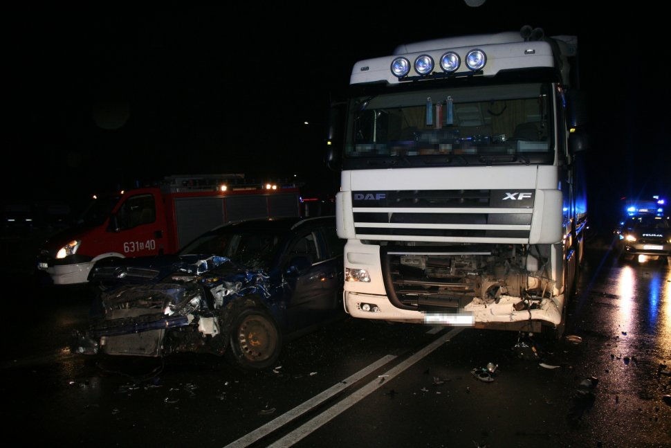 Na fotografii widać uszkodzony samochód ciężarowy, obok którego widać rozbity przód samochodu osobowego. W tle fotografii widać policyjny radiowóz