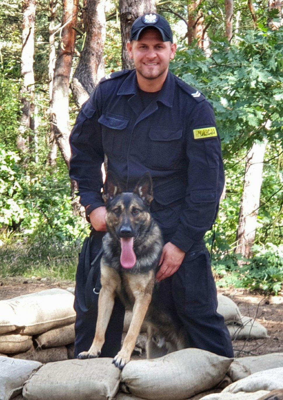 Na zdjęciu widać st. sierż. Kamila Kubicę, który trzyma na smyczy psa patrolowo-tropiącego. Policjant i pies znajdują się w okopie z workami piachu. 
