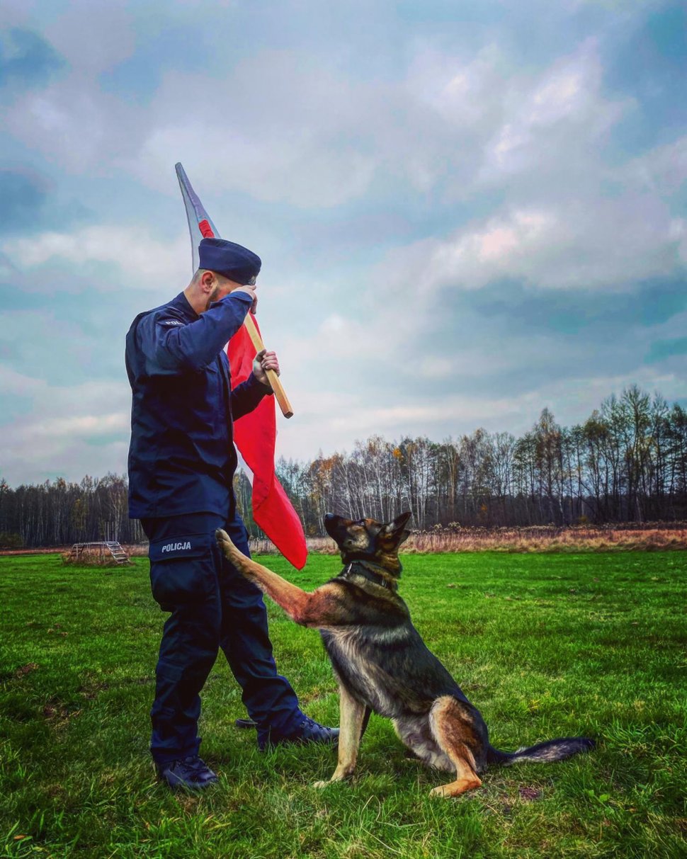 Na zdjęciu widać st. sierż. Kamila Kubicę, który trzyma w ręku biało-czerwoną flagę i oddaje honor. Obok niego znajduje się pies Otok, który opiera łapę na nodze policjanta.