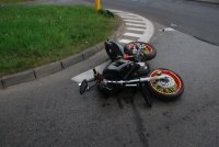 Zdjęcie przedstawia leżący na jezdni motocykl. Motocykl jest uszkodzony z uwagi na zderzenie z samochodem osobowym.