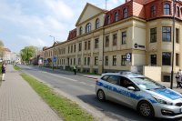 Fotografia kolorowa. Na zdjęciu widać budynek Komendy Powiatowej Policji w Tarnowskich Górach, obok której przejeżdża policyjny radiowóz