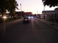 Na fotografii widać drogę, na której znajduje się samochód osobowy. Za samochodem widać dwa policyjne radiowozy z włączonymi sygnałami świetlnymi. Z prawej strony zdjęcia widać chodnik, na którym leży uszkodzony rower.