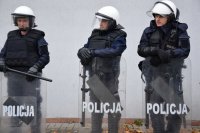 Na fotografii widać trzech policjantów, którzy są ubrani w kamizelki i hełmy. Policjanci mają przed sobą tarcze, na których widnieje napis POLICJA