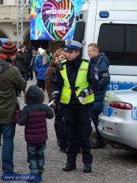 Na fotografii widać policjanta, przy którym stoi kobieta z dzieckiem. Na zdjęciu widać również inne osoby, dwa policyjne radiowozy oraz umieszczony na ścianie budynku znajdującej się za policjantem, plakat z napisem Wielka Orkiestra Świątecznej Pomocy