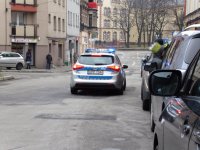 Na fotografii widać ulicę po której jedzie policyjny radiowóz. Z prawej strony zdjęcia widać zaparkowane przy ulicy pojazdy