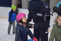 Na fotografii widać lodowisko, na którym znajdują się młodzi łyżwiarze. Dziewczynce, policjant zakłada na głowę kask narciarski, który wygrała w konkursie.