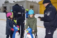 Na fotografii widać lodowisko, na którym znajdują się młodzi łyżwiarze. Dziewczynce, policjant zakłada na głowę kask narciarski, który wygrała w konkursie. Obok nich, znajduje się chłopiec i drugi policjant