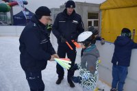 Na fotografii widać lodowisko, na którym znajdują się młodzi łyżwiarze. Jednemu z nich, policjant zapina na rękę odblaskową opaskę.