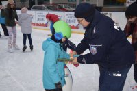 Na fotografii widać lodowisko, na którym znajdują się młodzi łyżwiarze. Jednemu z nich, policjant zapina na rękę odblaskową opaskę.