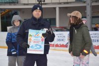 Na fotografii widać lodowisko, na którym stoi policjant. Mundurowy trzyma w ręku plakat z kampanii &quot;Kreci mnie bezpieczeństwo na stoku&quot;. obok niego widać młodych łyżwiarzy