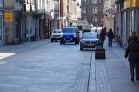 fotografia kolorowa: na zdjęciu widać ulicę, po której jedzie policyjny radiowóz. Obok niego znajdują się zaparkowane samochody oraz przemieszczające się osoby.