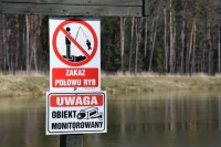 fotografia kolorowa: znak, na którym widnieje napis &quot;zakaz połowu ryb&quot; oraz &quot;obiekt monitorowany&quot;. W tle widać zalew