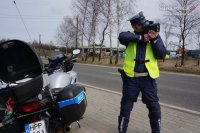 fotografia kolorowa: policjant przykłada do oka urządzenie służące do pomiaru prędkości pojazdów. ZX lewej strony zdjęcia widać zaparkowany policyjny motocykl.