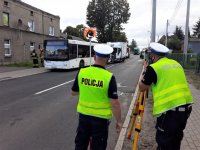 zdjęcie przedstawia dwóch policjantów stojących obok urządzenia służącego do wykonywania pomiarów. W tle widać uszkodzony autobus oraz ciężarówkę.
