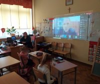 Na zdjęciu widać szkolną klasę. Przy biurku siedzi wychowawca, a na ekranie rzutnika wyświetla się wizerunek policjantki prowadzącej zajęcia online.