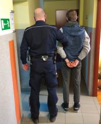 Zdjęcie przedstawia odwróconych tyłem: policjanta, który trzyma za rękę zatrzymanego mężczyznę. Sprawca ma założone kajdanki na ręce trzymane z tyłu.