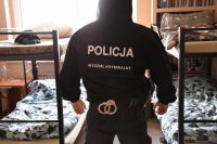 Zdjęcie kolorowe, na którym widać policjanta ubranego w czarną kurtkę z kapturem, na której jest napisane Policja Wydział Kryminalny. Policjant stoi w pomieszczeniu, w którym znajdują się dwa piętrowe łóżka.