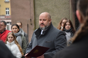 na zdjęciu przedstawiciel stowarzyszenia rodzin katyńskich, który w ręku trzyma bordową teczką z godłem polskim, za nim młodzież
