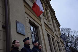 na zdjęciu policjanci i młodzież w trakcie warty pod pamiątkową tablicą wmurowaną w budynek komendy, nad nimi flaga polski