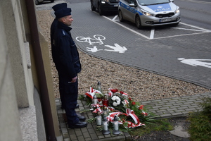 na zdjęciu policjanci pod tablicą, przed nimi złożone kwiaty i znicze, w oddali radiowóz