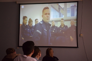 na zdjęciu ekran, na którym wyświetlany jest policjant, jego wizerunek obserwują uczniowie przed ekranem
