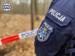 na zdjęciu fragment policyjnego munduru z naszywką oraz fragment lasu ogrodzony taśmą z napisem policja